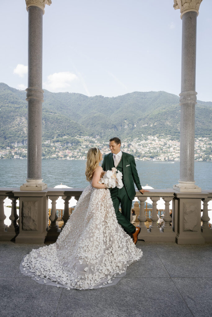 Bridal Couple facing each other, standing on the terrace of mandarin oriental, in the background you can see lake como. the bride is wearing a Oscar de la Renta dress with 3D flower applications, the groom is wearing a dark green Brioni suit