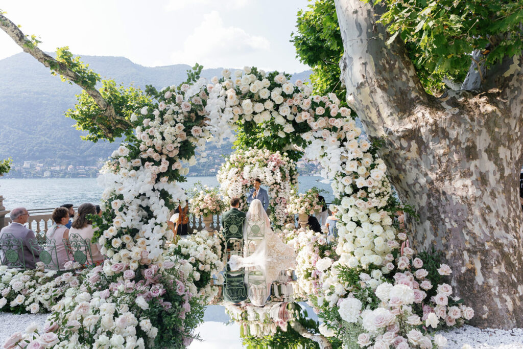 Wedding Ceremony with mirror aisle, multiple flower archs, flower meadows, the bridal couple sitting in front of their Guests facing Lake como