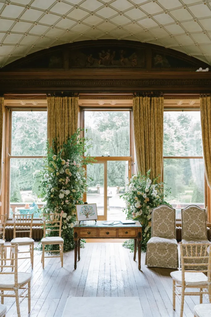 Kulisse für die Indoor-Trauung im Inneren von Castle Leslie mit üppigem Broken Arch Blumenbogen vor großen Fenstern