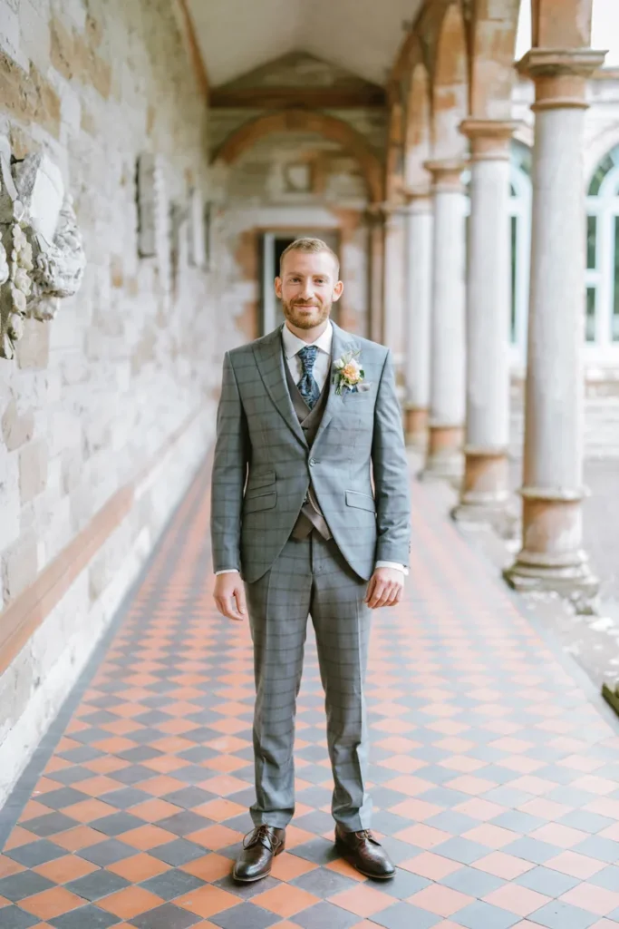 Smiling groom in a stylish three-piece suit standing under the stone arcades at Castle Leslie Estate