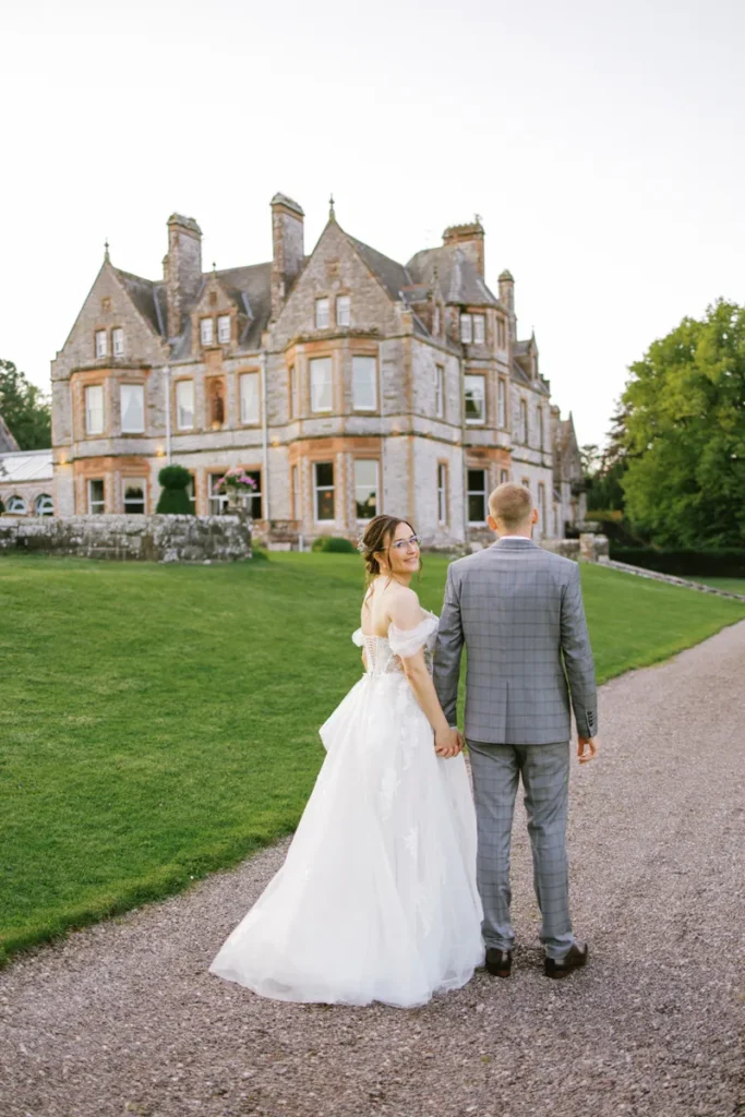 Braut blickt lächelnd über die Schulter, während das Paar zur Blauen Stunde auf Castle Leslie zugeht