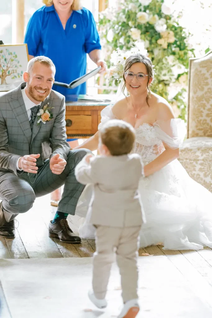 Bride and groom kneeling and smiling at the groom's young nephew as he delivers the wedding rings