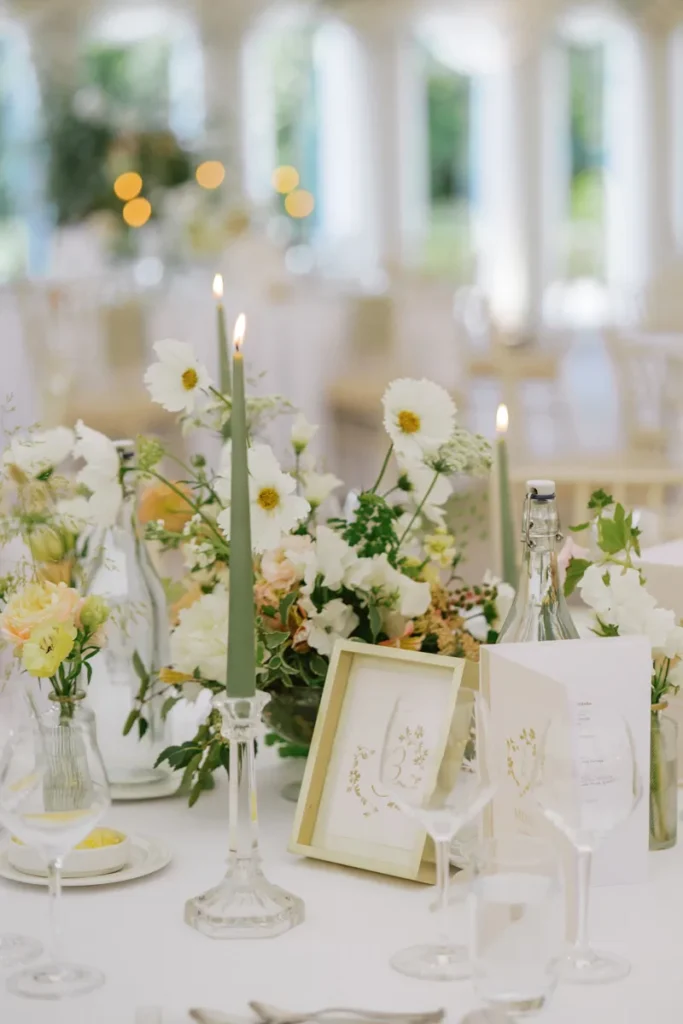 Close-up of a wedding table setting with lit sage green taper candles, fresh flowers, and a custom table number
